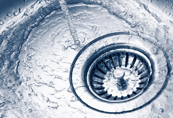 Close-up of water flowing from a faucet into a stainless steel sink, with droplets splashing around a metal drain. The image has a cool, blue-tinted tone and highlights quality plumbing Chicago professionals provide.