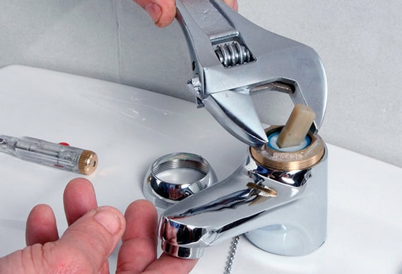A person in IL uses a wrench to repair a chrome bathroom faucet, with the handle and cover removed and a screwdriver nearby, demonstrating common DIY skills found among HVAC Chicago professionals.