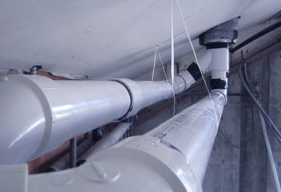 White PVC pipes are installed along a ceiling in an industrial space, supported by straps and brackets, with additional wiring and concrete walls visible—typical of plumbing Chicago utility installations.