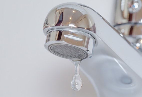 Close-up of a chrome bathroom faucet with a single droplet of water hanging from the spout, poised to fall, against a blurred white background—a simple scene highlighting the precision found in quality plumbing Chicago professionals deliver.