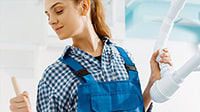 A woman wearing a blue checkered shirt and blue overalls holds a paint roller in one hand and a white ladder in the other, appearing to be renovating indoors—perhaps updating her IL home with new plumbing or HVAC Chicago services.