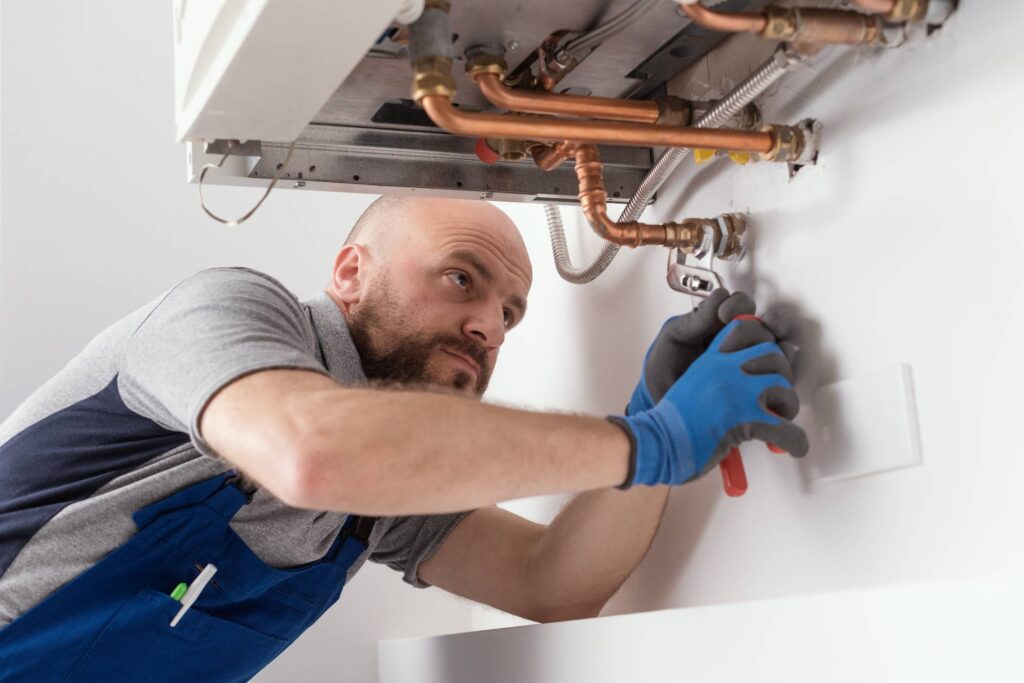 A plumber wearing blue gloves and overalls uses a wrench to work on pipes beneath a wall-mounted boiler or water heater. He appears focused on tightening or adjusting the fittings.