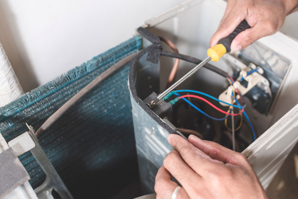 Close-up of a person’s hands using a screwdriver to repair or assemble the internal components of an air conditioning unit, with exposed wires and metal parts visible.